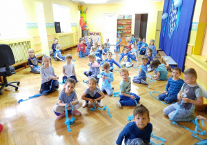 Dzieci siedzą na podłodze sali gimnastycznej trzymając niebieskie paski bibuły